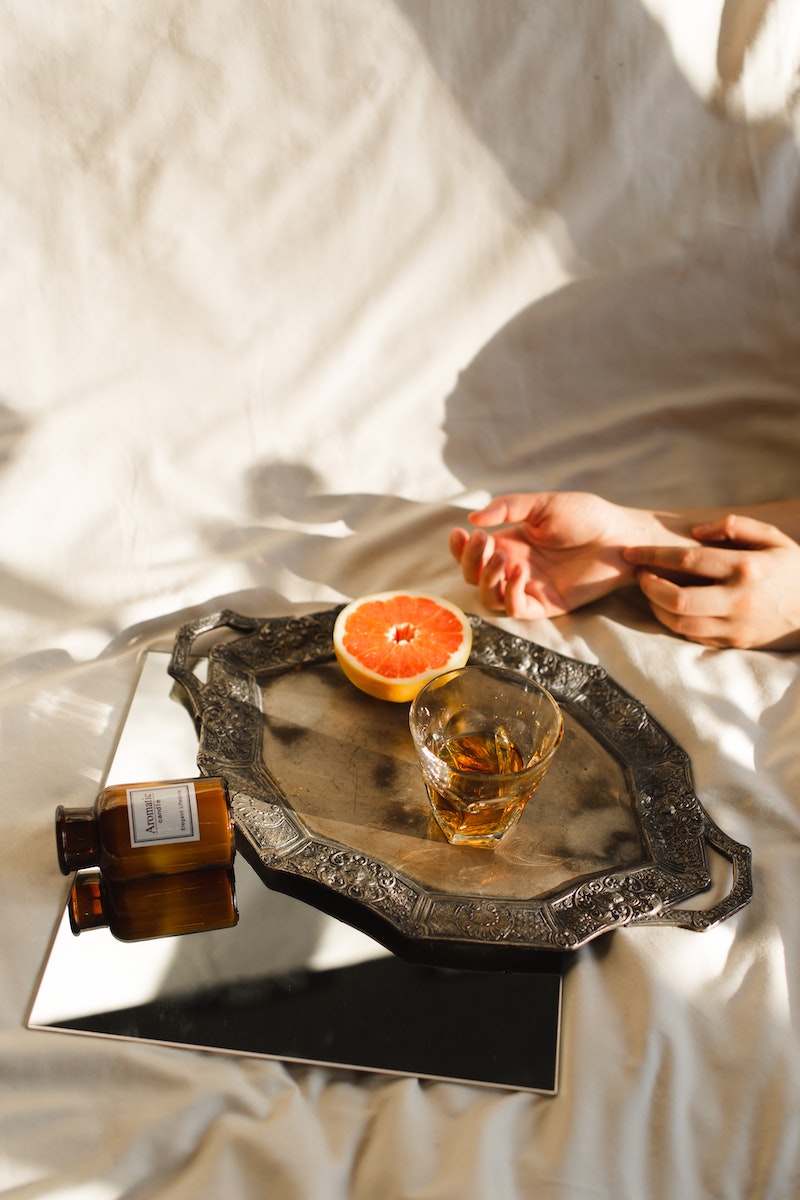 From above of crop anonymous female resting on crumpled bed sheet with ripe orange and alcoholic drink in glass on tray